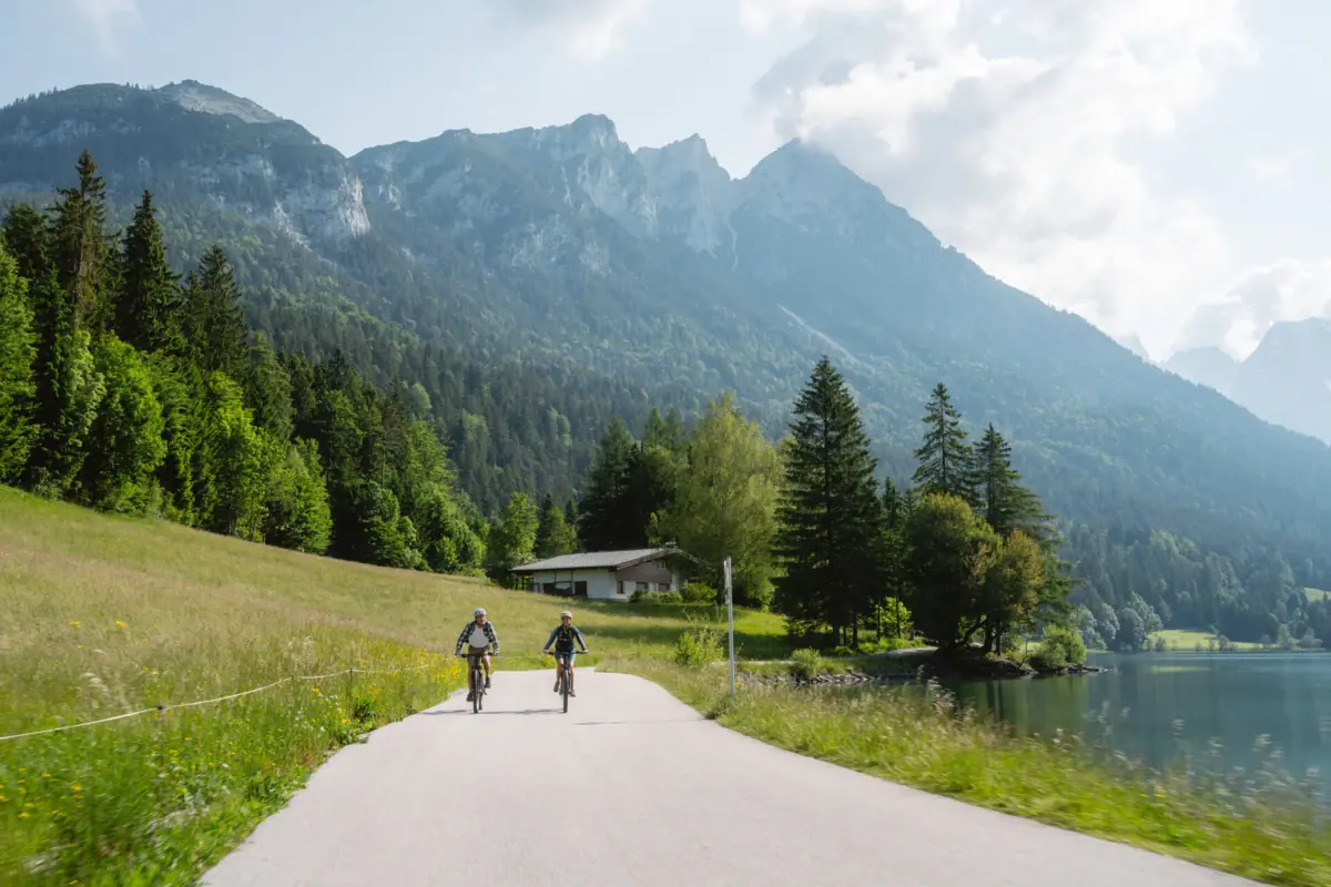 Cycle tour with mountains in the background Two people are riding bicycles on a road near a lake.