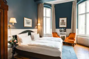 HENRI Hotel Berlin Boutique Zimmer Design Blau Stilvolles Hotelzimmer im HENRI Berlin mit dunkelblauen Wänden, hohen Decken, zwei orangenen Sesseln und einem eleganten Doppelbett im historischen Ambiente.