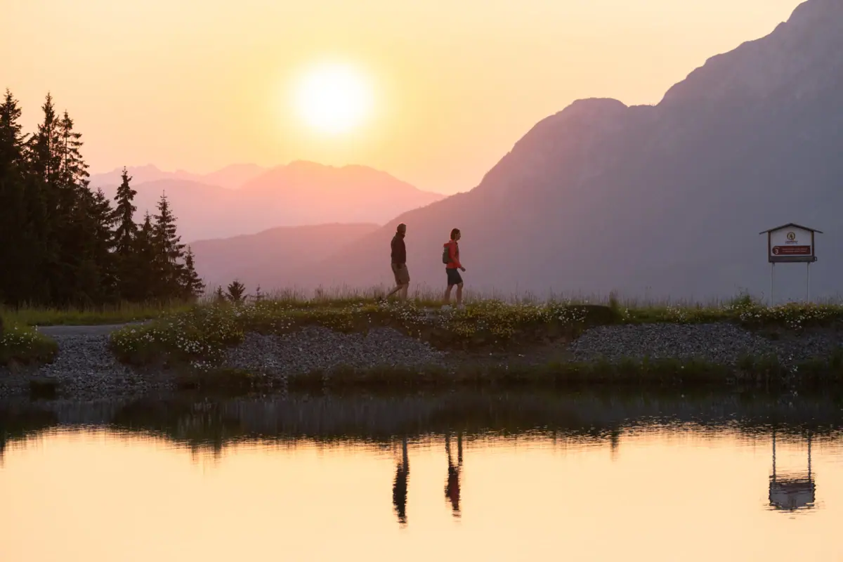 Sunset in front of a mountain lake Two people are walking on a hill by a lake.