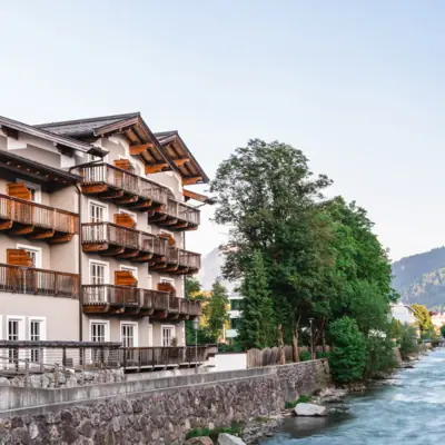 HENRI Hotel Kitzbühel Außenansicht Das HENRI Hotel Kitzbühel von außen neben einem Fluss und Bäumen.