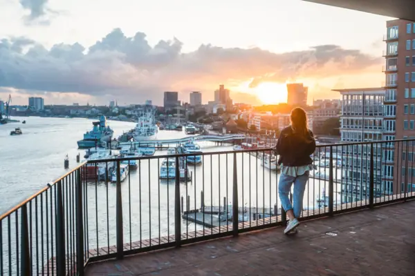 Hamburg Elphie A woman stands on a balcony and looks out over a body of water with boats in the background.