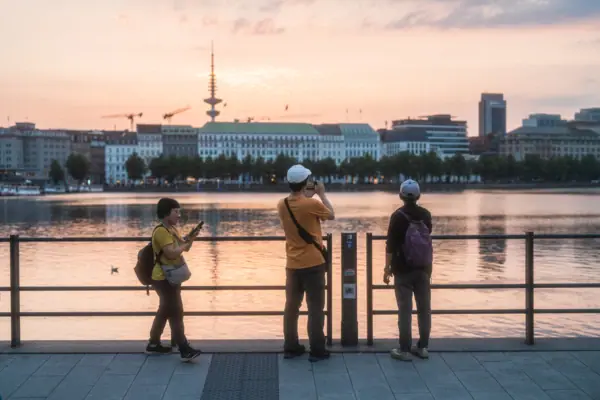 Alster A group of people are standing on a pavement next to a body of water.