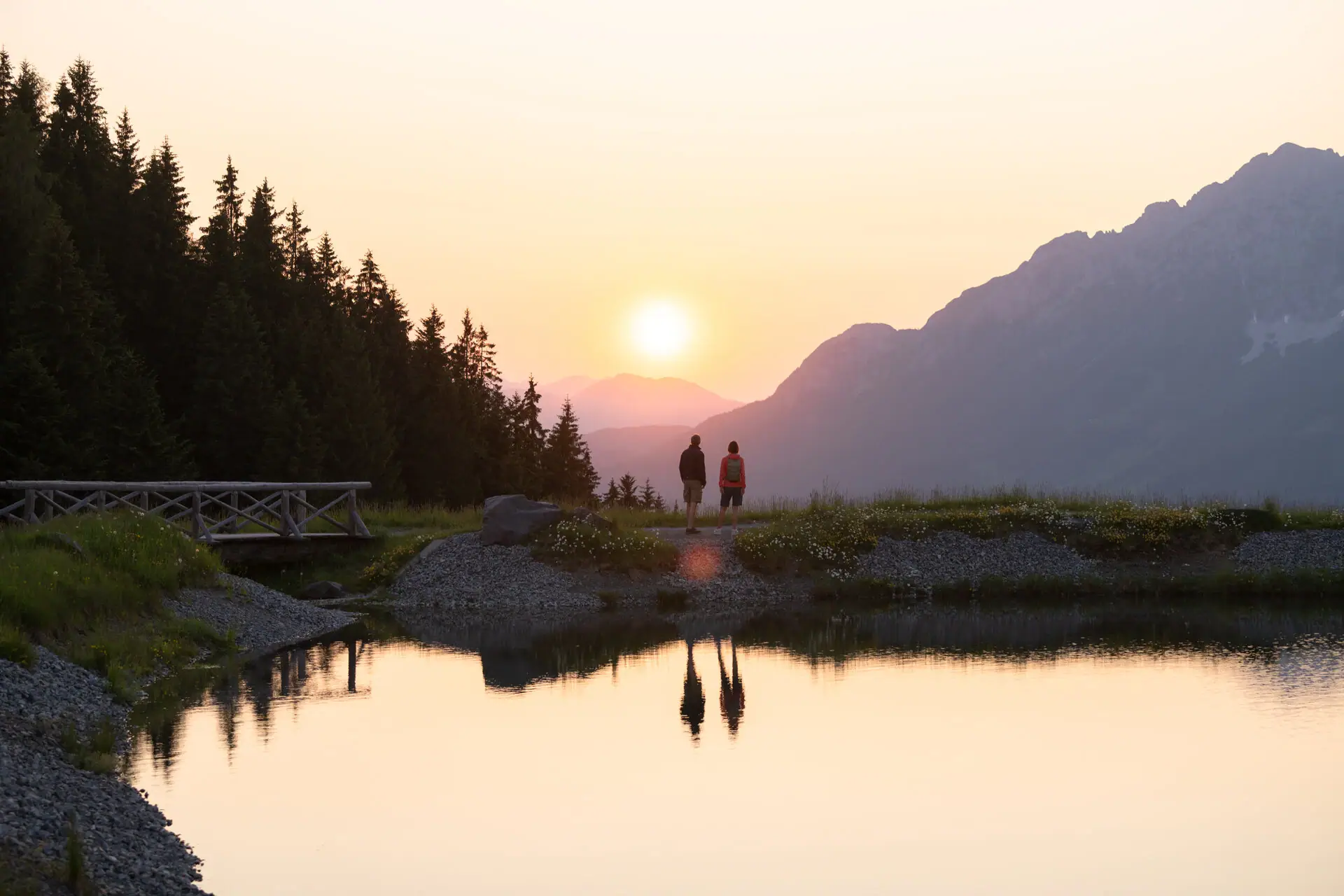 Mountain lake in Kitzbühel Two people walk along the rocky shore of a lake, with trees and mountains in the background.